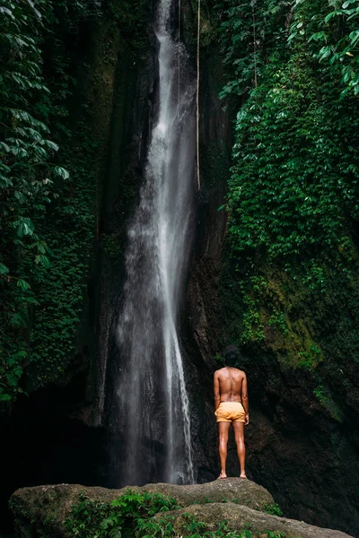 A dark-skinned man in yellow shorts at a waterfall on the island of Bali. A man travels the world. Man at the waterfall, view from the back. Travel to Bali Indonesia. Man on the background of nature