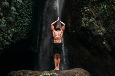 Atletik yapılı bir adam yoga yapar. Sağlıklı bir yaşam tarzı. Vücudun konsantrasyonu. Bir erkek doğada yoga yapar. Bali 'de yoga yapan bir adam. Bir adam doğada meditasyon yapar. Şelalede meditasyon
