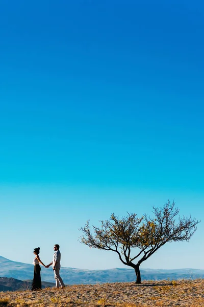 Um casal solitário junto a uma árvore ao pôr-do-sol. Um casal apaixonado conhece o pôr-do-sol. Um homem e uma mulher encontram o amanhecer nas montanhas. Amantes ao amanhecer. Sigam-me. Casal no fundo da natureza — Fotografia de Stock