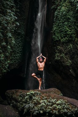 Atletik yapılı bir adam yoga yapar. Sağlıklı bir yaşam tarzı. Vücudun konsantrasyonu. Bir erkek doğada yoga yapar. Bali 'de yoga yapan bir adam. Bir adam doğada meditasyon yapar. Şelalede meditasyon