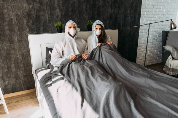 夫婦は家で隔離されている フードとフェイスマスクを持つ白い保護衣装の素敵なカップルは ベッドの上に家に座っています コロナウイルスの隔離Covid 19のために単離された — ストック写真
