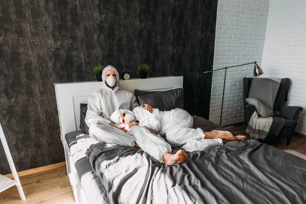 夫婦は家で隔離されている フードとフェイスマスクを持つ白い保護衣装の素敵なカップルは ベッドの上に家に座っています コロナウイルスの隔離Covid 19のために単離された — ストック写真