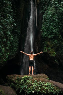 Back view of a man standing by waterfalls with arms outstretched. Man at waterfall raising his hands in feeling closer to nature. Man at the waterfall. Travel to Bali. Man on the background of nature. With copy space