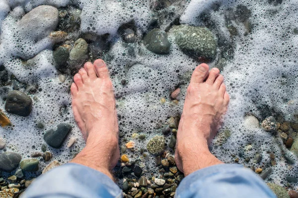 Голые ноги человека, который стоит на галечном пляже рядом с морской волной. Пены для воды . — стоковое фото