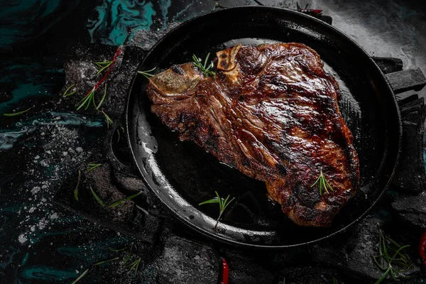 Fried meat steak on a black background of charcoal. Cooked juicy steak with smoke on the coals with rosemary. Grilled delicious marinated steak on coals, closeup photo. — Stock Photo, Image