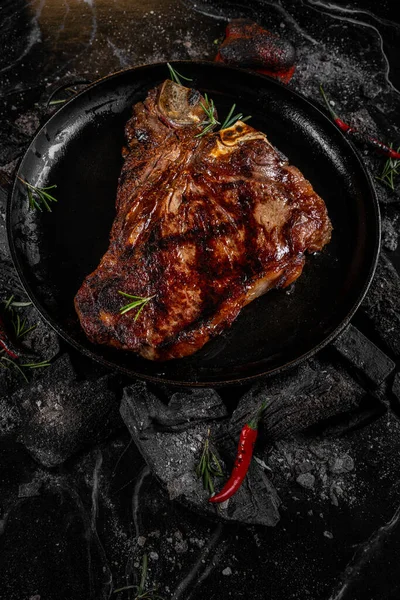 Fried meat steak on a black background of charcoal. Cooked juicy steak with smoke on the coals with rosemary. Grilled delicious marinated steak on coals, closeup photo. — Stock Photo, Image