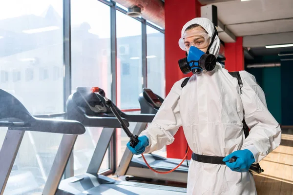 コロナウイルス流行中の混雑した場所での清掃と消毒ジムの清掃と消毒感染予防と流行の制御 保護スーツとマスクとスプレーバッグ — ストック写真