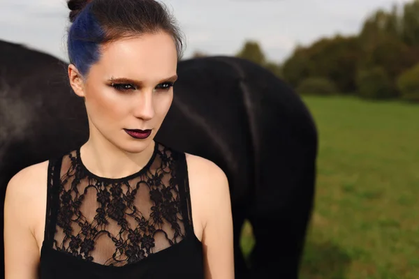 Elegantes Mädchen mit langen Haaren und blauen Haarsträhnen in schwarzem Kleid, das auf einem Feld mit schwarzem Pferd steht. — Stockfoto