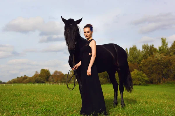 우아한 여자 긴 머리와 파란색 검은 검은 말과 함께 필드에 서 있는 검은 드레스에 머리의 자물쇠. — 스톡 사진