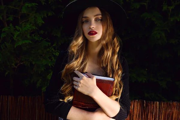 年轻漂亮的女孩，长长的卷发，她手里拿着本书戴黑帽子的红色嘴唇 — 图库照片