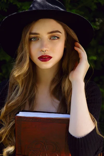 Young beautiful girl with long curly hair and red lips in black hat with book in her hands — Stock Photo, Image