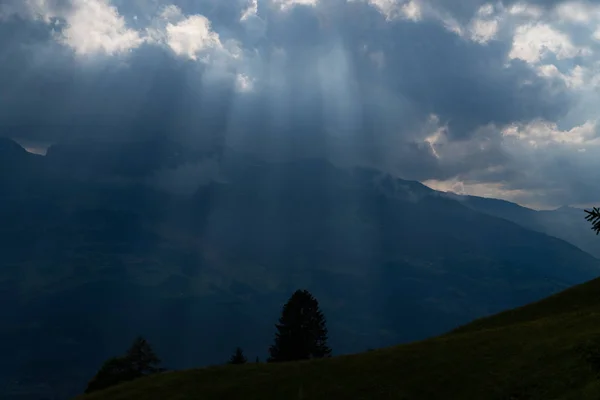 Sun rays shining trough the clouds in the mountains, a hill and trees in front — 스톡 사진