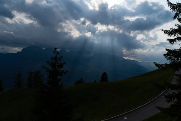 Raios de sol brilhando através das nuvens nas montanhas, uma colina, uma rua e árvores na frente — Fotografia de Stock