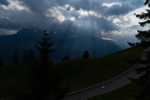 Raios de sol brilhando através das nuvens nas montanhas, uma colina, uma rua e árvores na frente — Fotografia de Stock