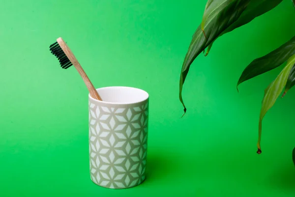One bamboo toothbrush in a mug with some leaves on green background — Stock Photo, Image