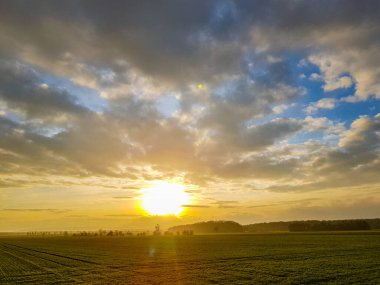 Bulutlu gökyüzünde gün batımı yeşil bir tarlanın üzerinde, dağların arkasında.