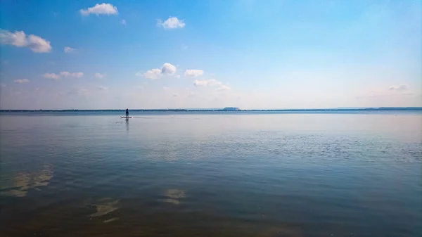 Un gran lago plano con el agua azul th y el cielo azul nublado y un remo de pie en la distancia — Foto de Stock