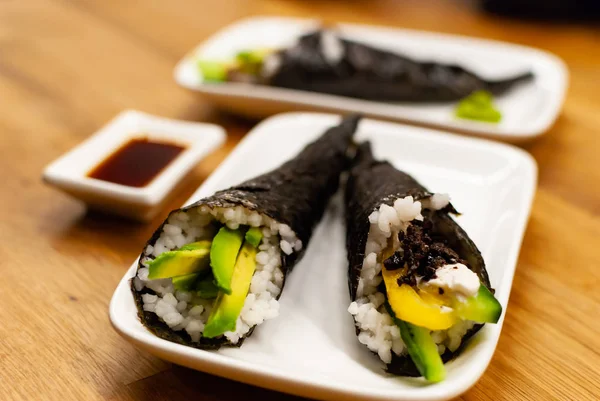 Temaki-Sushi vegan de casă umplut cu avocado pe o farfurie mică pe o masă de lemn cu sos de soia și wasabi — Fotografie, imagine de stoc
