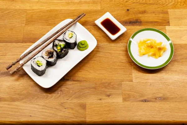 Role vegane de sushi de casă umplute cu avocado pe o farfurie mică pe o masă de lemn cu sos de soia, wasabi și ghimbir — Fotografie, imagine de stoc