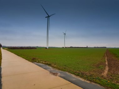 Bir bisiklet yolu, yeşil bir tarla ve mavi gökyüzünden önce arkada rüzgar değirmenleri