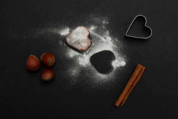 Angled view of a cookie with sugar powder, nuts, a cinnamon roll and a heart shaped baking form on black background — Stock Photo, Image