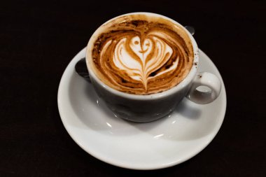 Bir fincan kapuçino. Süt köpüğünün içinde bir çiçek. Siyah arka planda.