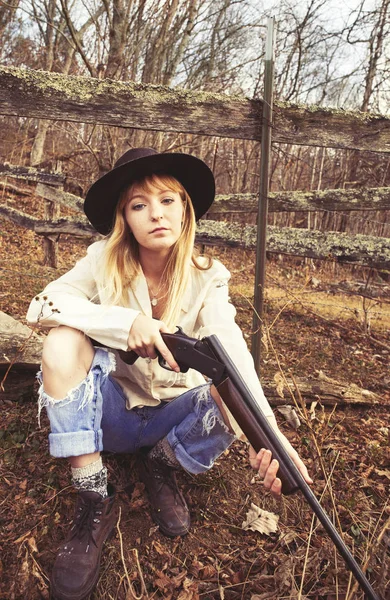 Mujer rubia joven con sombrero sentado en el bosque con un arma — Foto de Stock