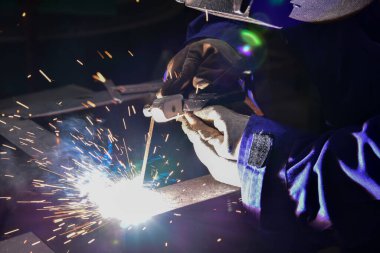 Kaynakçı, kalkanlı metal yay kaynaklama (SMAW) ile iş parçalarını birleştiriyor. Mavi üniformalı kaynakçı, güvenlik ayakkabıları, deri eldivenler, kaynak maskesi. Oturuyor ve kaynak yapıyor..