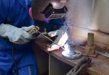 Kaynakçı, kalkanlı metal yay kaynaklama (SMAW) ile iş parçalarını birleştiriyor. Mavi üniformalı kaynakçı, güvenlik ayakkabıları, deri eldivenler, kaynak maskesi. Oturuyor ve kaynak yapıyor..