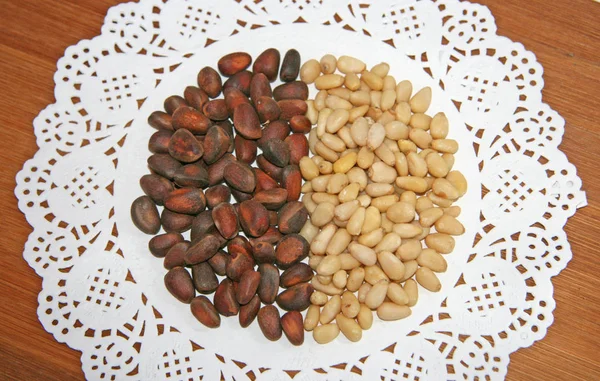 Pine nuts are laid out in the form of a circle. Next to brown and beige on white. A white napkin with a pattern on it are nuts.  Nuts in the shell and peeled from a pine cone. Cedar tree seeds are laid out next to each other. Collection of nuts.