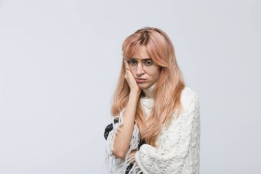 Studio portrait of nervous girl in white sweater and glasses, presses hand to face, doesn't know what to do, looking down/found out what happened, bad news, nervous feeling, experiences of fear.