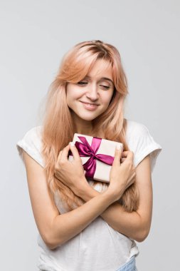Vertical studio portrait of cute blonde girl in white t-shirt presses the gift to the chest/beautiful girl received a gift on Valentine's Day/pleasant surprise, gift in box, joyful glee, lovers day.