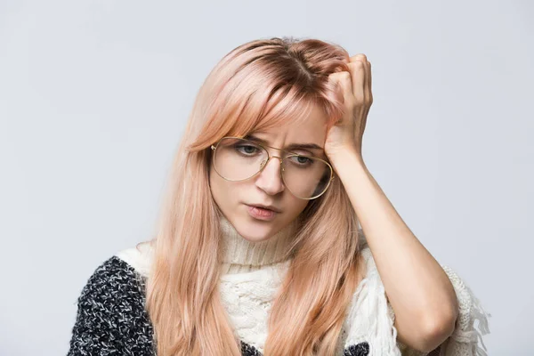Indoor portrait of pretty female in white sweater with eyewear doubting in correctness of taken decision, presses hand to head/plans something on coming weekends/thoughtful woman remember something.