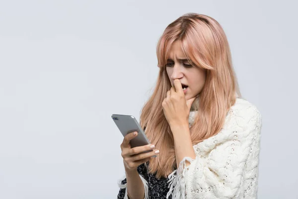 Studio portrait of blonde teen in white sweater waiting for a response sms, gnawing nails, looking at the phone/ found out what happened, bad news, nervous feeling, experiences of fear/people concept