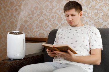 Genç beyaz adam kitap okuyor. Yakınlarda beyaz, modern hava nemlendirici var. Erkeğe odaklan. Aroma yağı yayıcı. Rahat bir günlük hayat. Hava saflığı ve sağlık hizmeti kavramı.