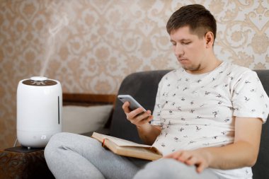 Genç beyaz adam kanepede oturup internette sörf yapıyor. Yakınlarda beyaz, modern hava nemlendirici var. Erkeğe odaklan. Rahat bir günlük hayat. Hava saflığı ve sağlık hizmeti kavramı.