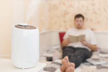 Beyaz nemlendirici buhar yayıyor. Oturma odasındaki kuru havanın nemlendirilmesi. Buhar üzerine seçici odaklanma. Arka planda kitap okuyan genç bir adam. Hava saflığı ve sağlık hizmeti konsepti. İç mimari