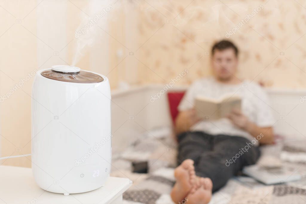 White humidifier spreading steam. Humidification of the dry air in living room. Selective focus on vapor. On the background young man reading book. Air purity and healthcare concept. Home interior
