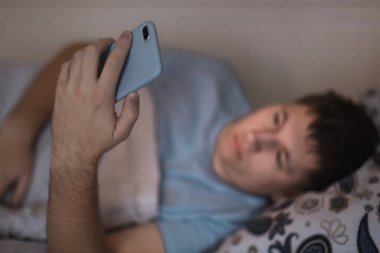 Uykulu yorgun erkek gece geç saatte yatakta uyandı ve ağda sörf yaptı. Sosyal medya bağımlılığı, cep telefonuna bağımlılık gözler üzerinde olumsuz etki, uykusuzluk kavramı. Akıllı telefona seçici odaklanma.