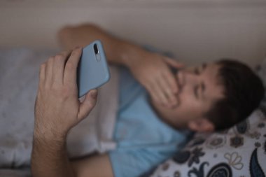Uykulu esneyen erkek gece geç saatte yatakta. İnternette sörf yapan bir adam uyuyamaz. Sosyal medya bağımlılığı, cep telefonuna bağımlılık. Uykusuzluk ve uykusuzluk kavramı. Eldeki seçici odaklanma.