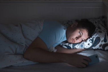 Uykulu erkek gece geç saatte yatakta internette sörf yapıyor, sohbet ediyor ve flört ediyor, uykulu yorgun, sosyal medya bağımlısı, cep telefonuna bağımlı, gözler üzerinde olumsuz etki, uykusuzluk kavramı.