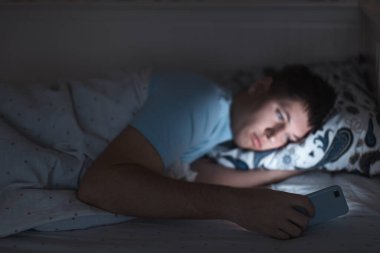 Uykulu yorgun erkek gece geç saatte yatakta uyandı ve ağda sörf yaptı. Sosyal medya bağımlılığı, cep telefonuna bağımlılık gözler üzerinde olumsuz etki, uykusuzluk kavramı. Akıllı telefona seçici odaklanma.