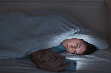 Uykulu depresif genç internette cep telefonuyla karanlıkta yatakta sörf yapıyor. Web bağımlılığı, akıllı telefon bağımlılığı, internet tuzağı. Uykusuzluk, nomofobi, uyku bozukluğu.