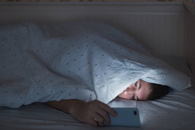 Uykulu yorgun bir adamın portresi battaniyenin altında uzanıyor ve gece yarısı akıllı telefon kullanıyor, uyuyamıyor. Uykusuzluk, nomofobi, uyku bozukluğu konsepti. Bir cep telefonuna bağımlılık. Yumuşak odak.