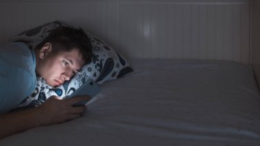 Uykulu erkek gece geç saatte yatakta uyanıyor ve internette sörf yapıyor ve sohbet ediyor. Sosyal medya bağımlılığı, cep telefonuna bağımlılık, gözler üzerinde olumsuz etki. Uykusuzluk ve uykusuzluk kavramı. Boşluğu kopyala.