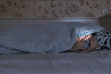 Yatakta battaniyenin altında yatan ve gece yarısı akıllı telefon kullanan genç, uykulu adamın portresi. Uykusuzluk, nomofobi, uyku bozukluğu konsepti. Cep telefonuna ve internet tuzağına bağımlılık.