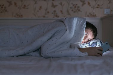 Battaniyenin altındaki genç beyaz adam akıllı telefonunu kullanarak internette sörf yapıyor. Sosyal medya bağımlılığı, cep telefonuna bağımlılık. Uykusuzluk ve uykusuzluk kavramı. Karamsar ışığı olan karanlık oda..