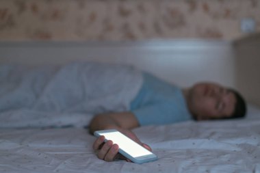 Genç adam yorgunluk sonucu elinde akıllı telefon ile uykuya daldı. Birikmiş yorgunluk, yeterli uyku ve bir cep telefonuna bağımlı olmama. Sağlık hizmeti konsepti. Telefona seçici odaklanma.