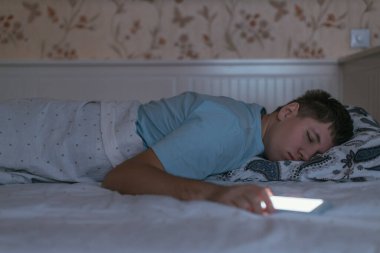 Genç giyinmiş adam elinde akıllı telefonuyla uyuyakaldı. Mavi tişörtlü bir erkek cep telefonunu çok kullanır ve uyku bozukluğundan muzdariptir. Sosyal medya ve internet bağımlılığı. Sağlık hizmeti kavramı.