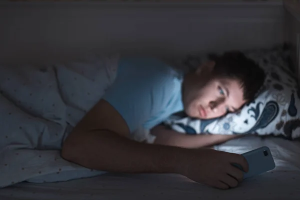 Uykulu yorgun erkek gece geç saatte yatakta uyandı ve ağda sörf yaptı. Sosyal medya bağımlılığı, cep telefonuna bağımlılık gözler üzerinde olumsuz etki, uykusuzluk kavramı. Akıllı telefona seçici odaklanma.